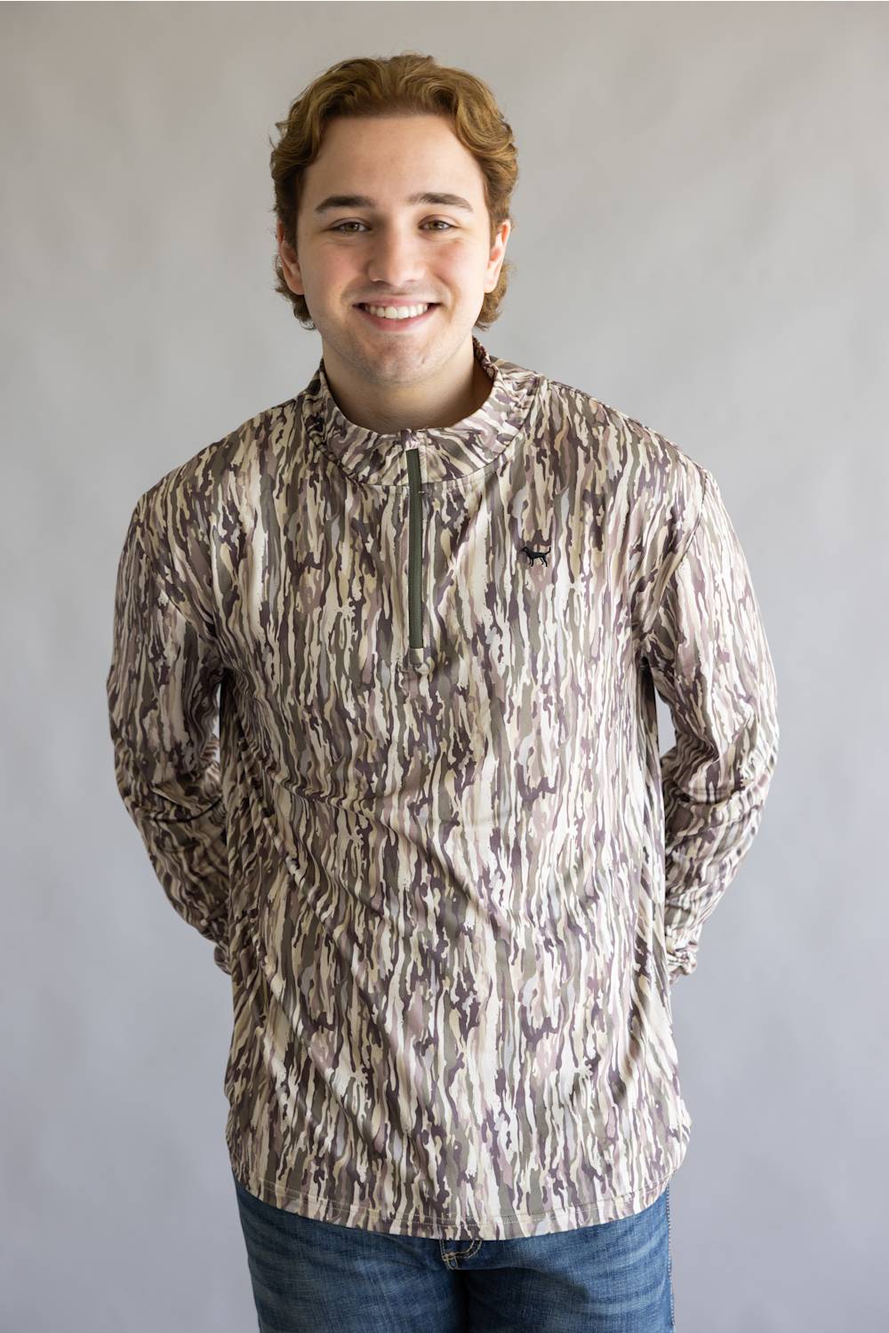 A smiling young man wearing a long-sleeve quarter-zip pullover jacket with a brown, beige, and gray bark-like camouflage pattern, paired with blue jeans, standing against a neutral gray studio background.