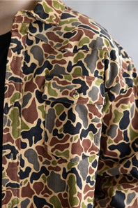 A close-up of a camouflage button-up shirt with a chest pocket, featuring a pattern of brown, green, black, and beige shapes.