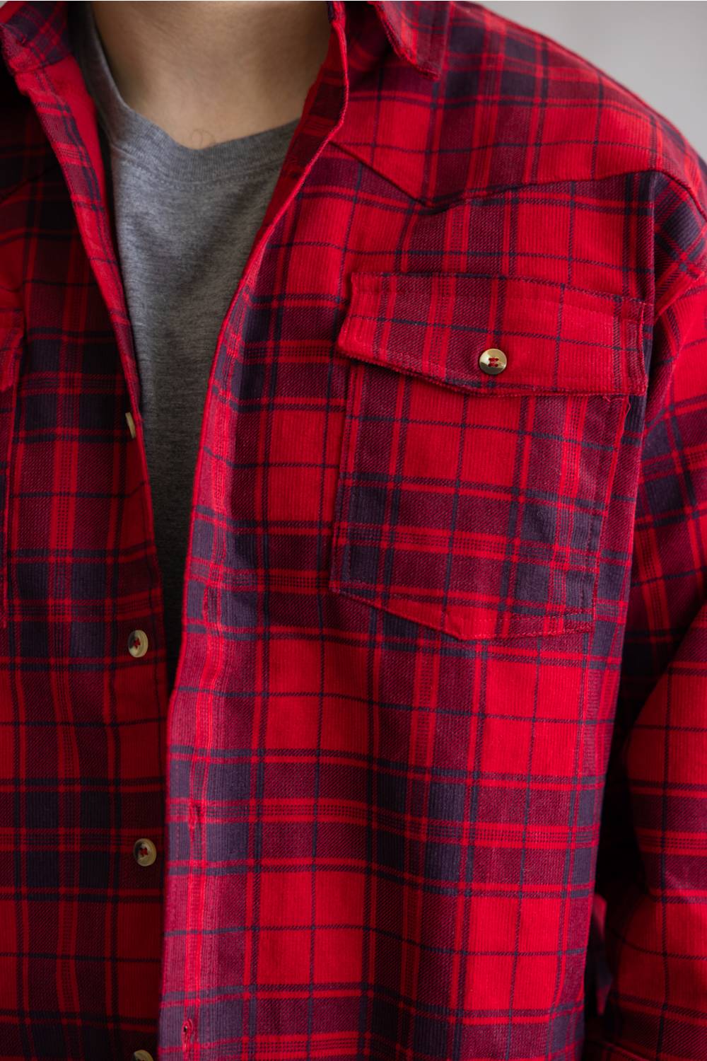 A close-up of a red and navy blue plaid flannel shirt worn open over a gray t-shirt, showing the chest pocket with a snap button.