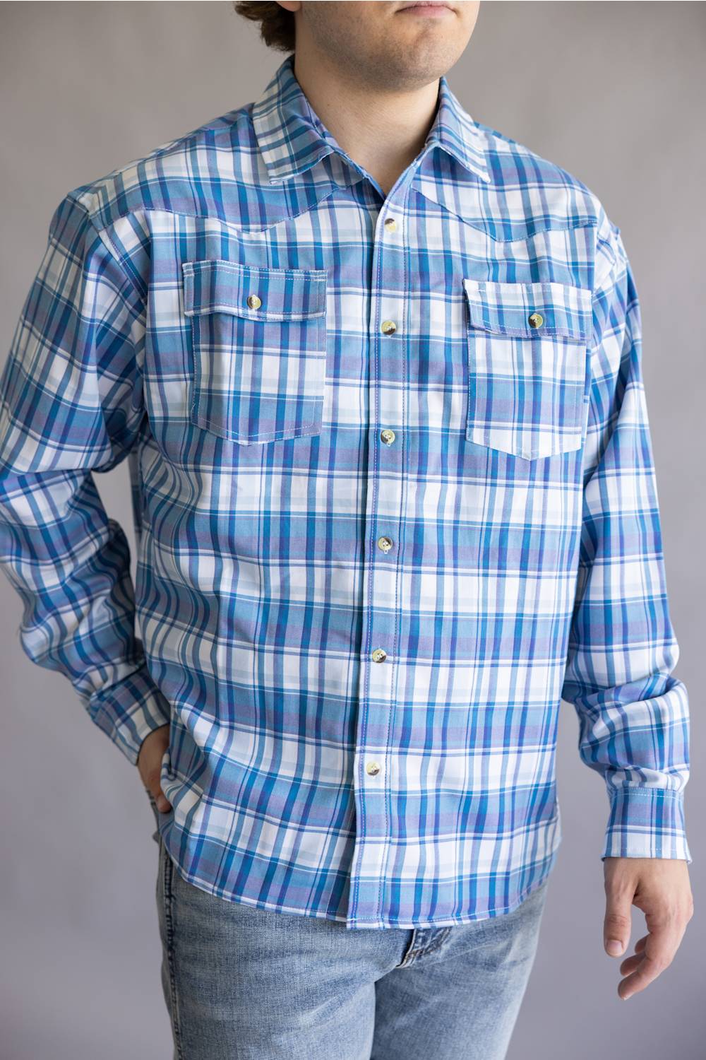 A man wearing a blue and white plaid long-sleeve button-up shirt with two chest pockets, paired with light-wash jeans, against a neutral gray background.
