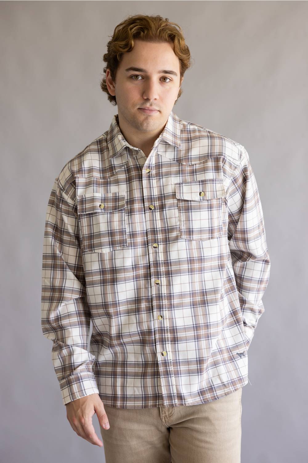 A man wearing a long-sleeve plaid button-up shirt in beige, brown, and white, paired with khaki pants, standing against a neutral gray studio background.