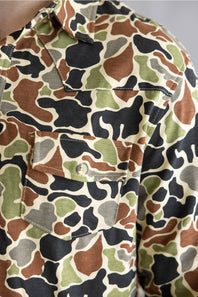 Close-up of a camouflage button-up shirt with a chest pocket and button closure, featuring a multi-color camo pattern in green, brown, black, and beige.