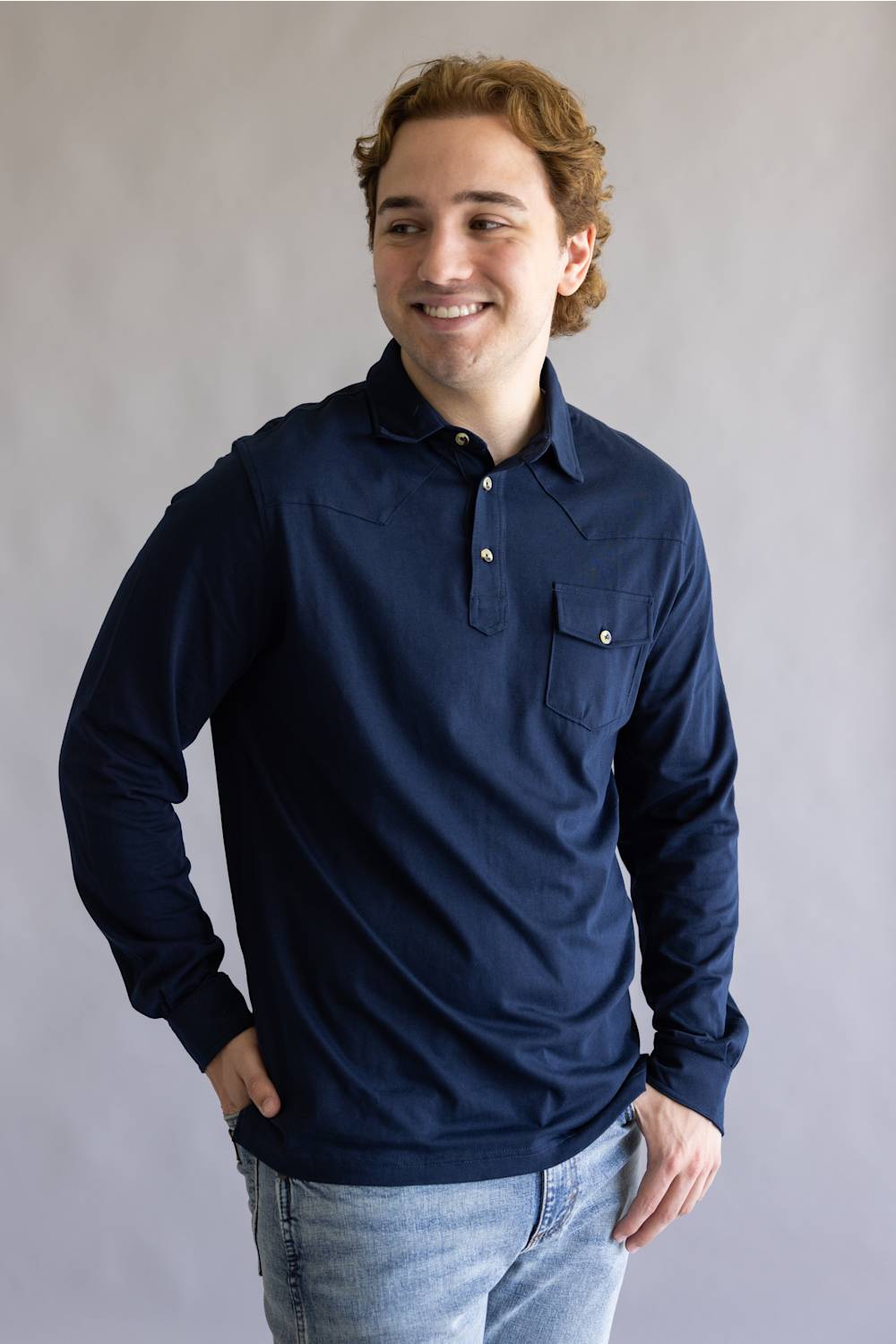 A smiling young man with light brown hair wearing a navy blue long-sleeve polo shirt and light wash jeans, standing against a neutral gray studio background.
