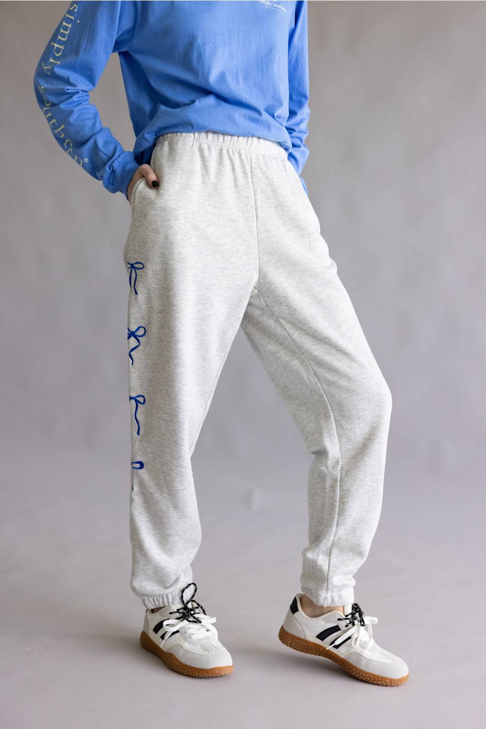 A person wearing light heather gray jogger sweatpants with blue embroidered ribbon details down the side, paired with a blue sweatshirt and white sneakers, shown from the chest down against a neutral gray studio background.