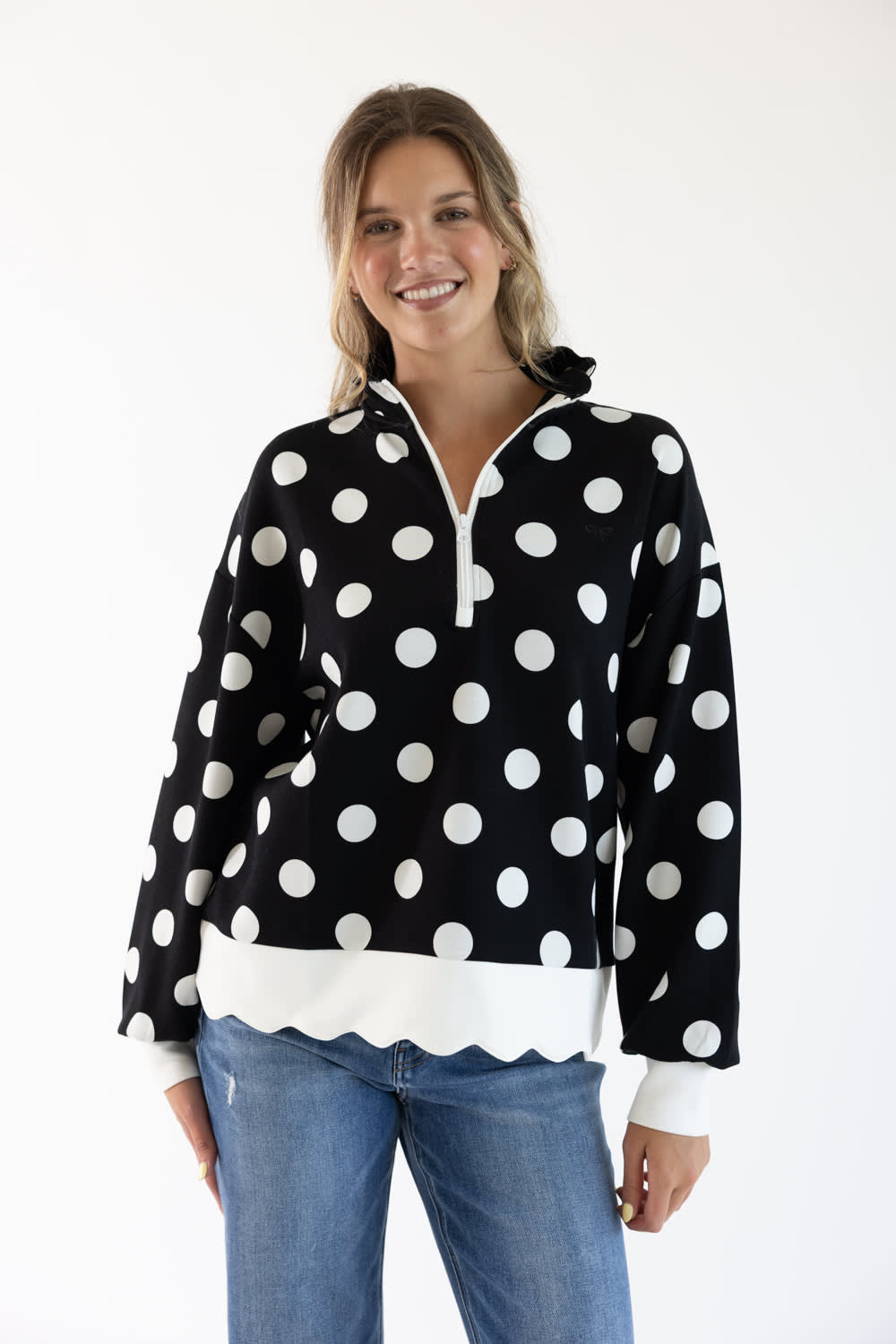 A woman modeling a black long-sleeve half-zip pullover with large white polka dots and a white scalloped hem, paired with blue jeans against a plain white background.
