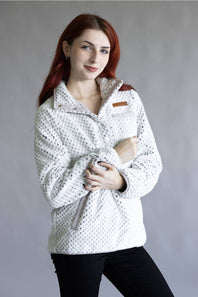 A woman with red hair wearing a white textured fleece pullover jacket with a subtle black pattern, black pants, and a small brown shoulder patch, posed against a neutral gray studio background.