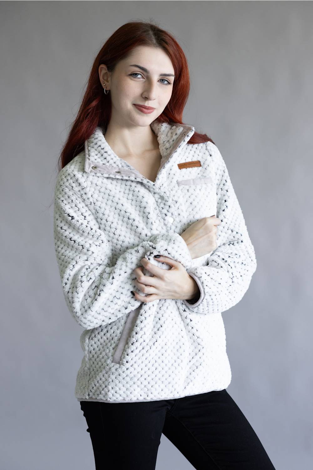 A woman with red hair wearing a white textured fleece pullover jacket with a subtle black pattern, black pants, and a small brown shoulder patch, posed against a neutral gray studio background.