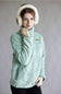 A woman wearing a light mint green textured fleece pullover jacket with a small brown chest patch, paired with white fluffy earmuffs, against a plain gray studio background.