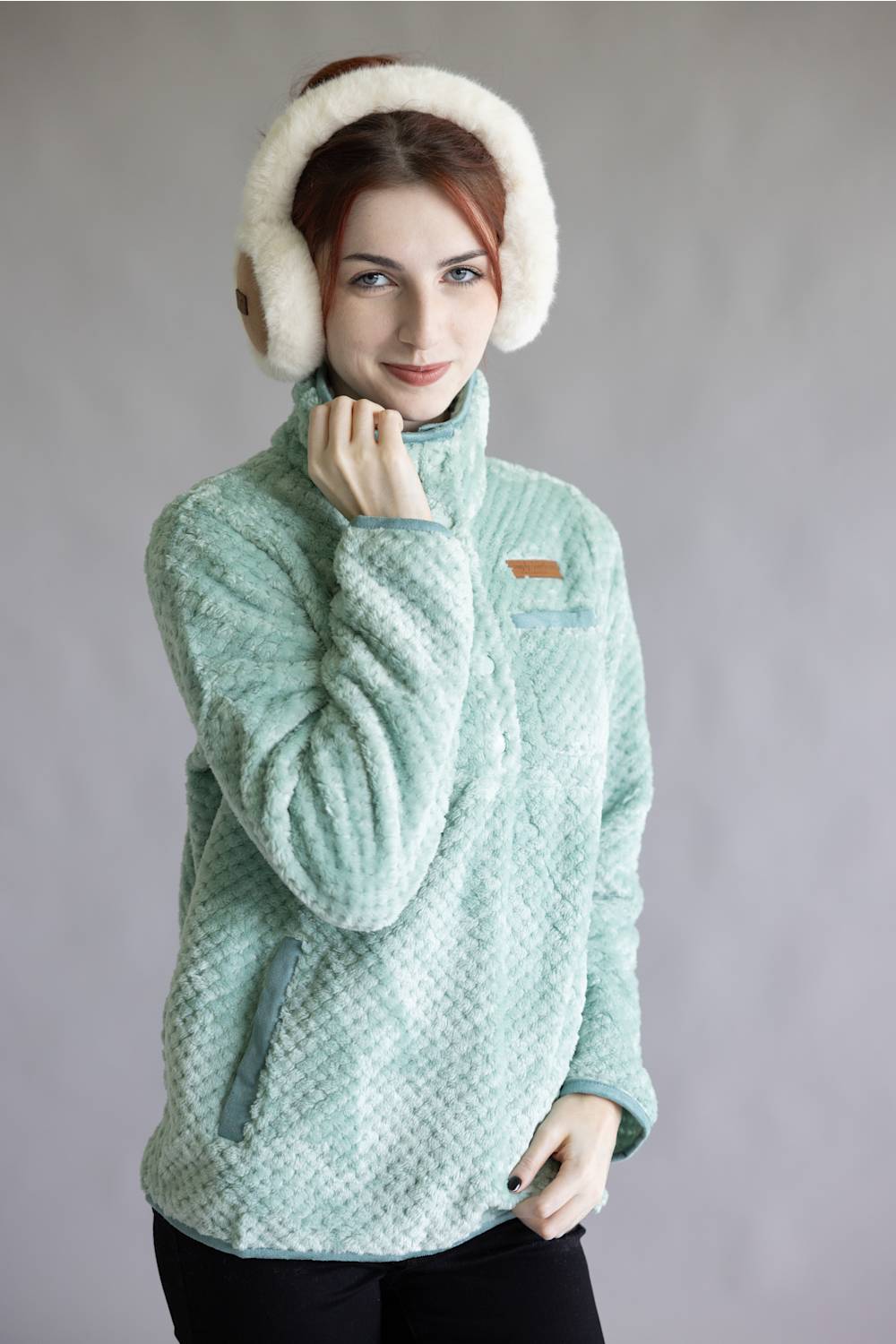 A woman wearing a light mint green textured fleece pullover jacket with a small brown chest patch, paired with white fluffy earmuffs, against a plain gray studio background.