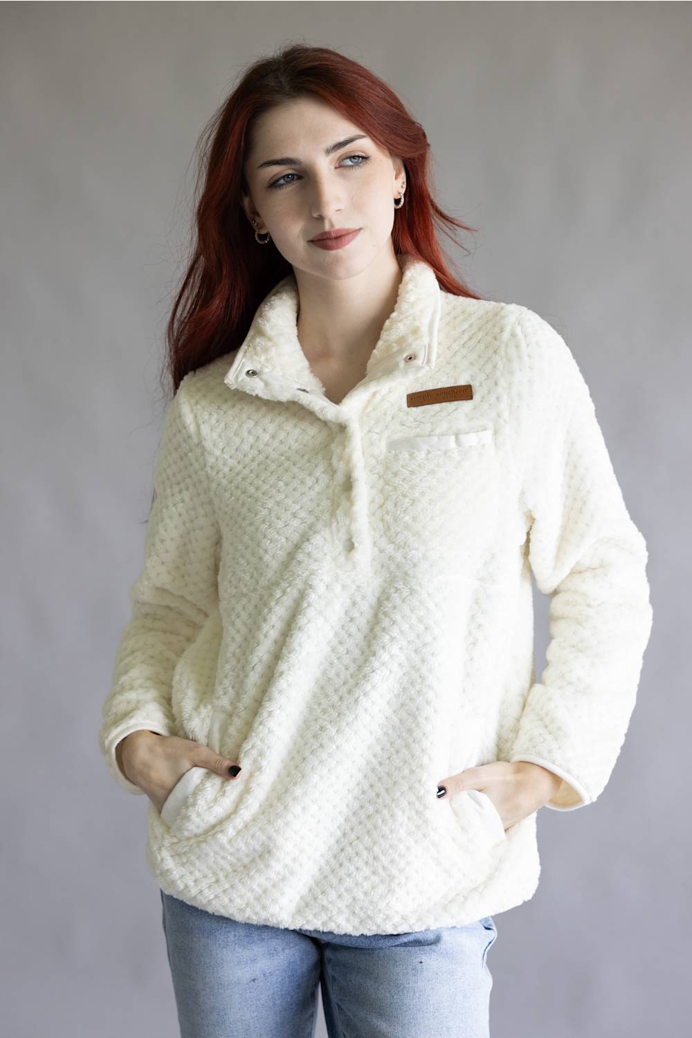 A person wearing a cream-colored textured fleece pullover jacket with a half-zip collar, a chest pocket, and a brown rectangular patch on the upper chest, paired with blue jeans against a neutral gray background.
