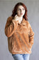 A woman with red hair wearing a textured, warm brown fleece pullover jacket and blue jeans, posing against a neutral gray studio background.
