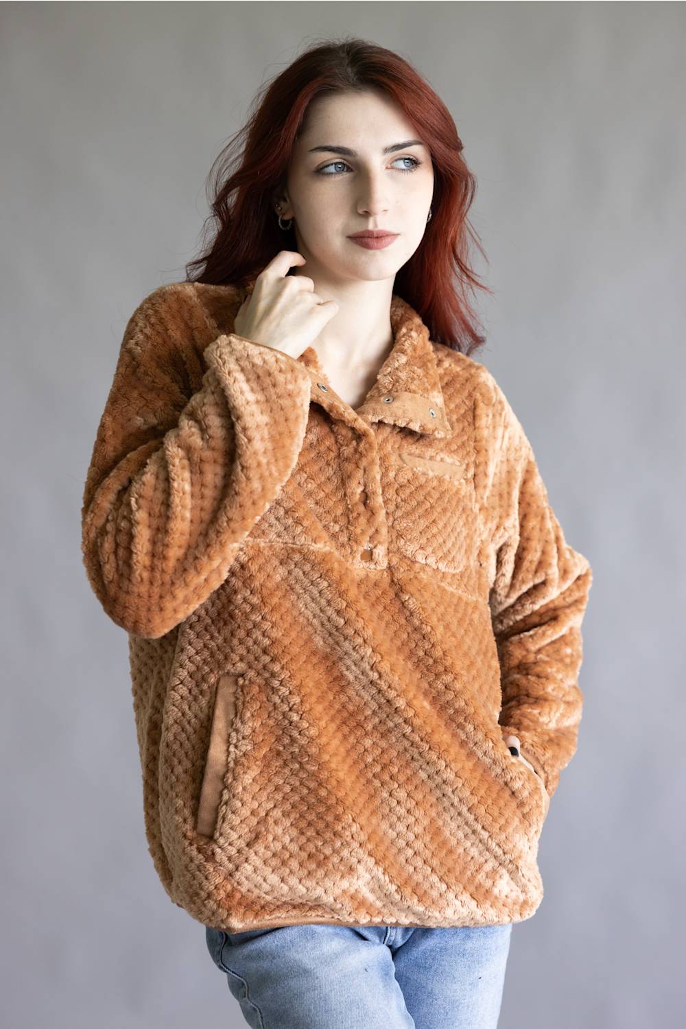 A woman with red hair wearing a textured, warm brown fleece pullover jacket and blue jeans, posing against a neutral gray studio background.