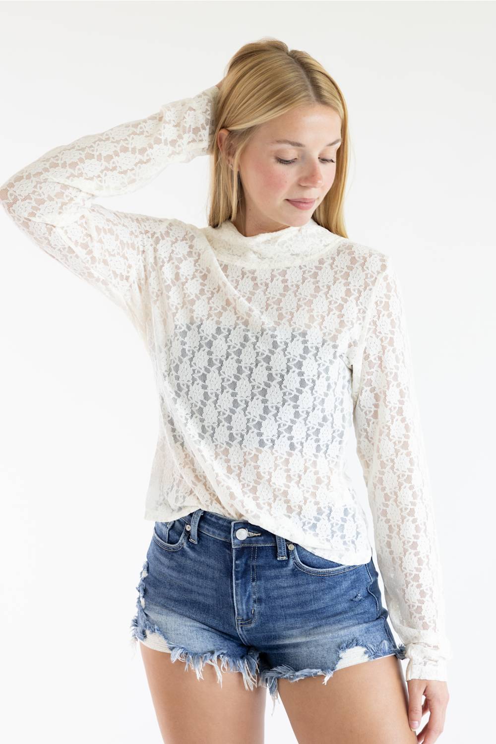 A woman wearing a white lace mock-neck long-sleeve top and distressed denim shorts, posing against a solid white background.