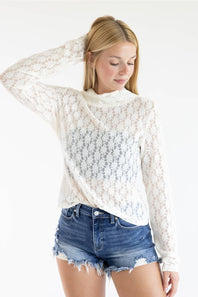 A woman wearing a white lace mock-neck long-sleeve top and distressed denim shorts, posing against a solid white background.