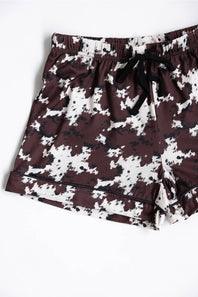 A pair of youth girls shorts featuring a dark brown and white cow pattern, with a black drawstring at the elastic waistband, laid flat against a white background.