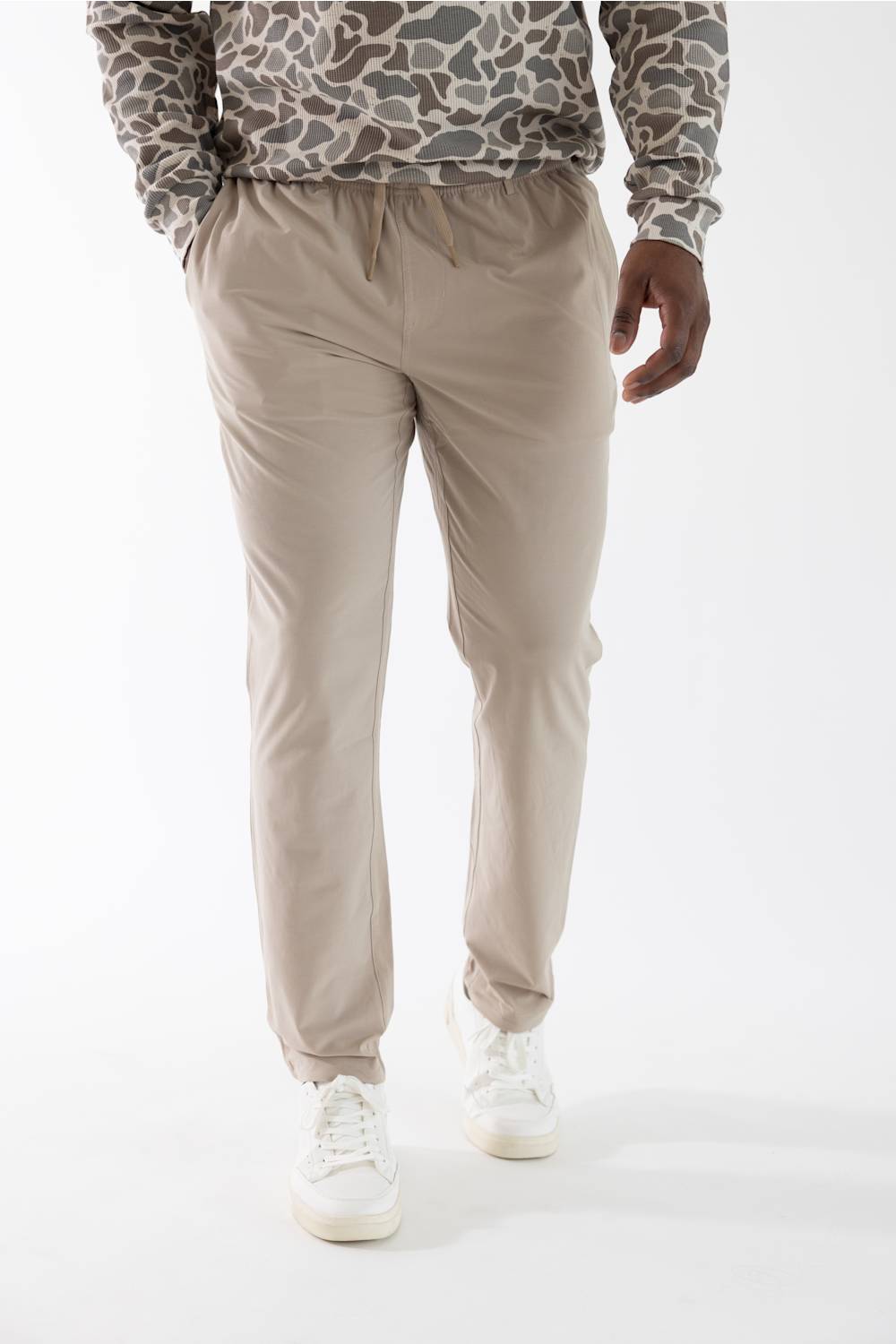 A person wearing beige drawstring jogger-style pants, a gray and brown camouflage hoodie, and white sneakers, photographed against a clean white studio background.