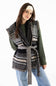 A woman modeling a black and cream geometric patterned knit wrap vest with a matching tie belt, worn over an olive green sweater and blue jeans against a plain white background.