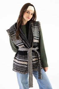 A woman modeling a black and cream geometric patterned knit wrap vest with a matching tie belt, worn over an olive green sweater and blue jeans against a plain white background.