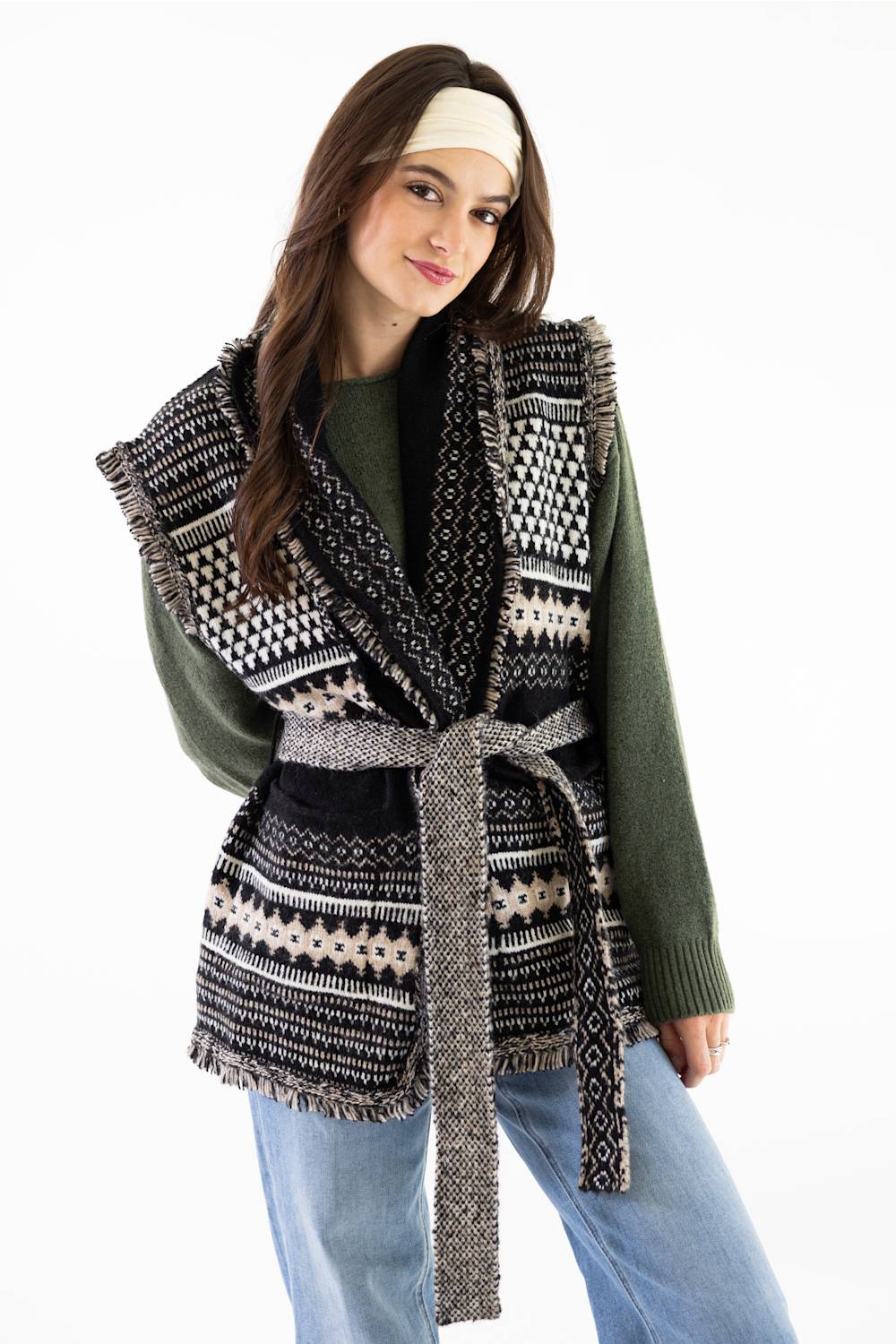 A woman modeling a black and cream geometric patterned knit wrap vest with a matching tie belt, worn over an olive green sweater and blue jeans against a plain white background.