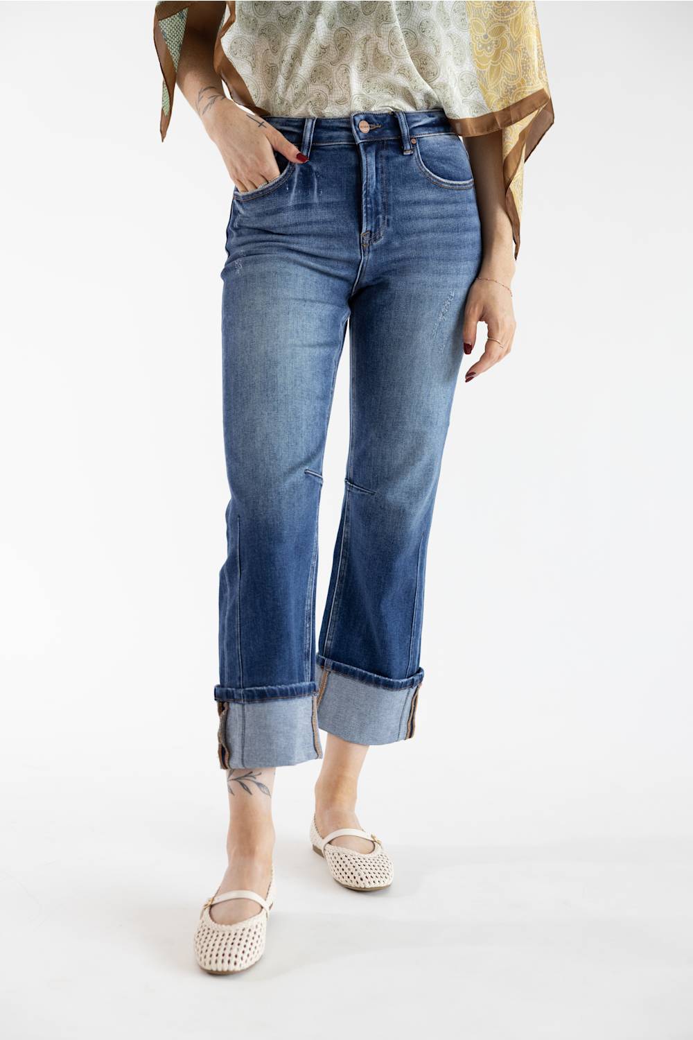 A model wearing a pair of medium-wash blue cropped jeans with rolled cuffs, paired with a patterned top and beige woven flats, shot against a plain white studio background.