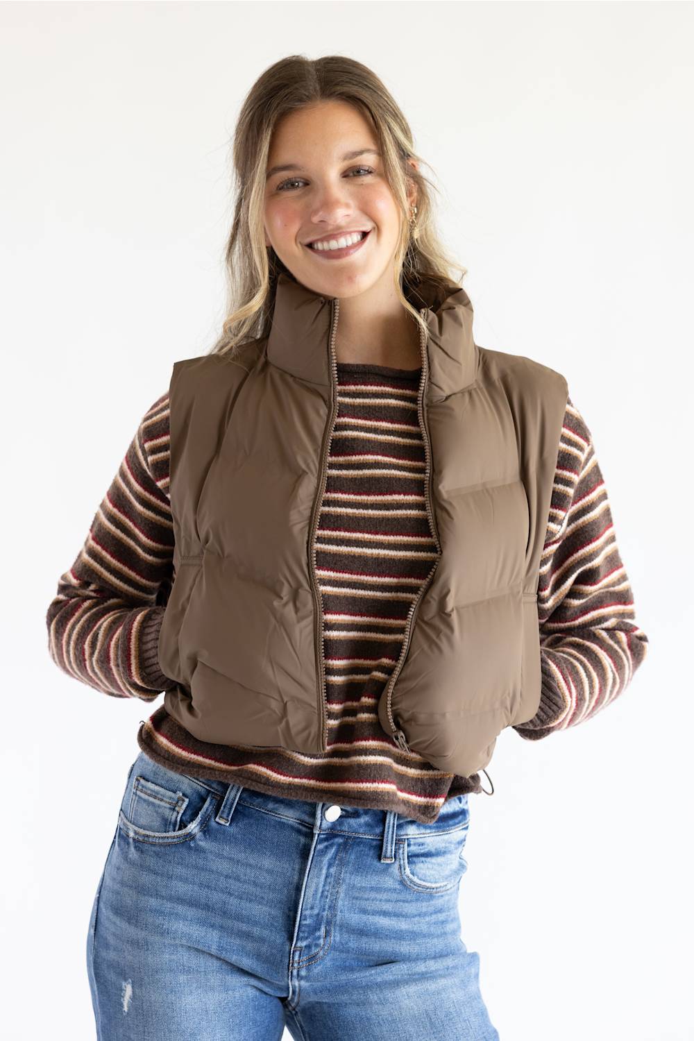 A smiling woman wearing a brown puffer vest over a brown and red striped long-sleeve shirt, paired with blue jeans, against a plain white background.