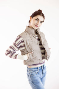 A woman modeling a sleeveless taupe puffer vest over a striped knit sweater and blue jeans, photographed against a plain white studio background.