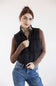 A woman wearing a black quilted puffer vest over a brown long-sleeve shirt and blue jeans, standing against a plain light gray studio background.