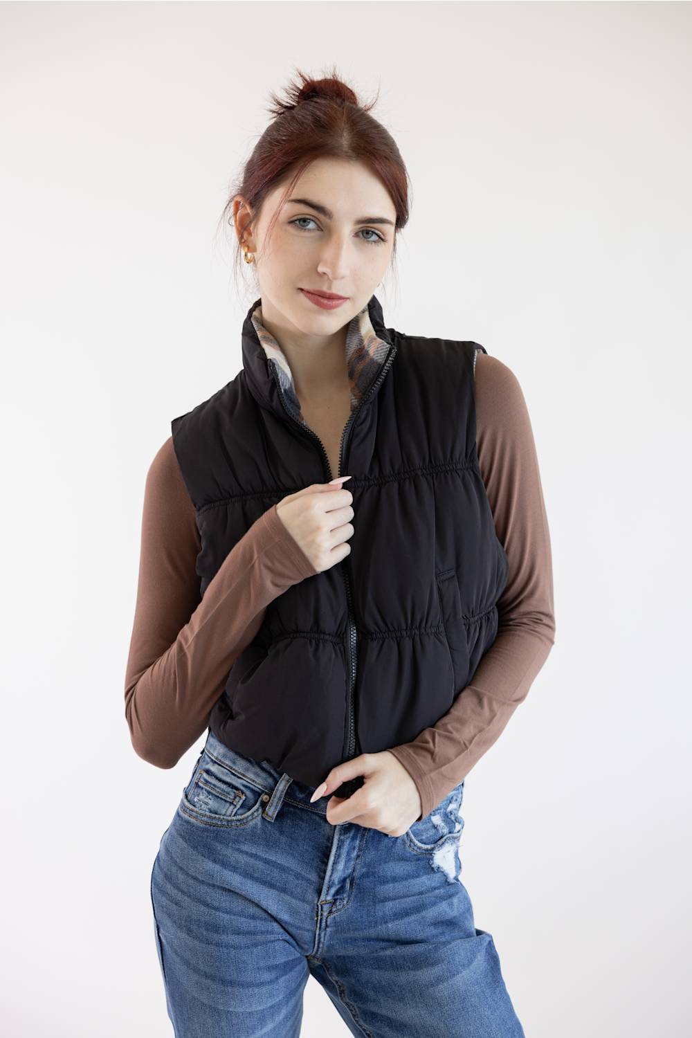 A woman wearing a black quilted puffer vest over a brown long-sleeve shirt and blue jeans, standing against a plain light gray studio background.