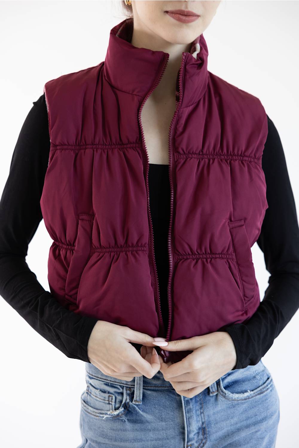 A woman wearing a sleeveless quilted puffer vest in deep burgundy over a black long-sleeve top and blue jeans, shown from the chin down against a plain white background.