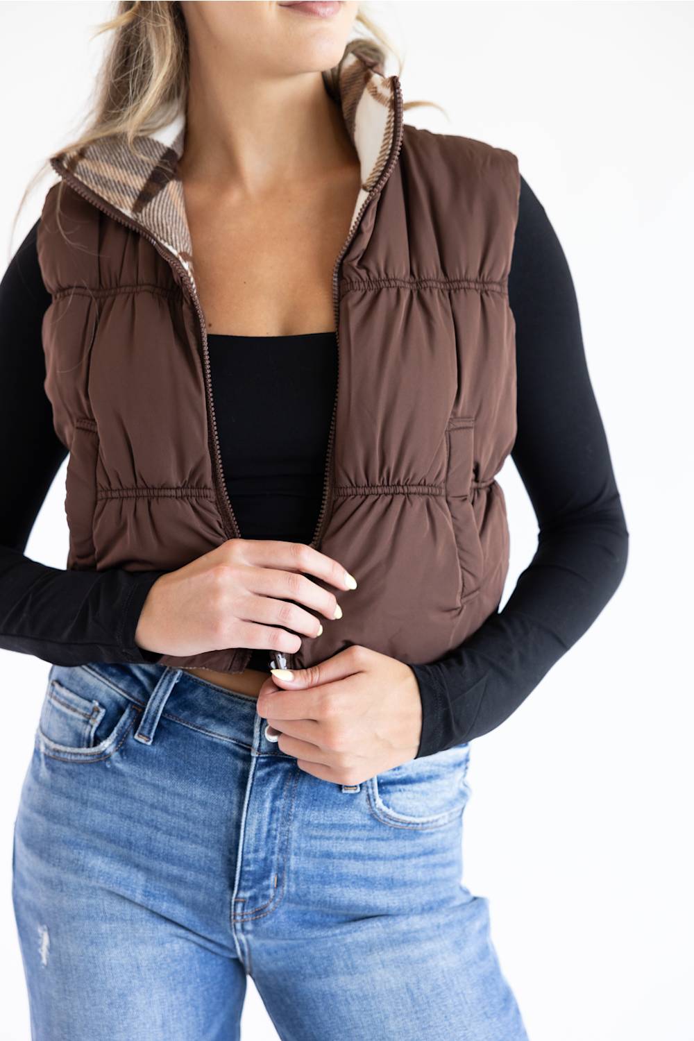 A woman wearing a brown puffer vest with a beige plaid lining over a black long-sleeve top and blue jeans, shown from the neck down against a white background.