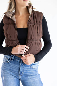 A woman wearing a brown puffer vest with a beige plaid lining over a black long-sleeve top and blue jeans, shown from the neck down against a white background.