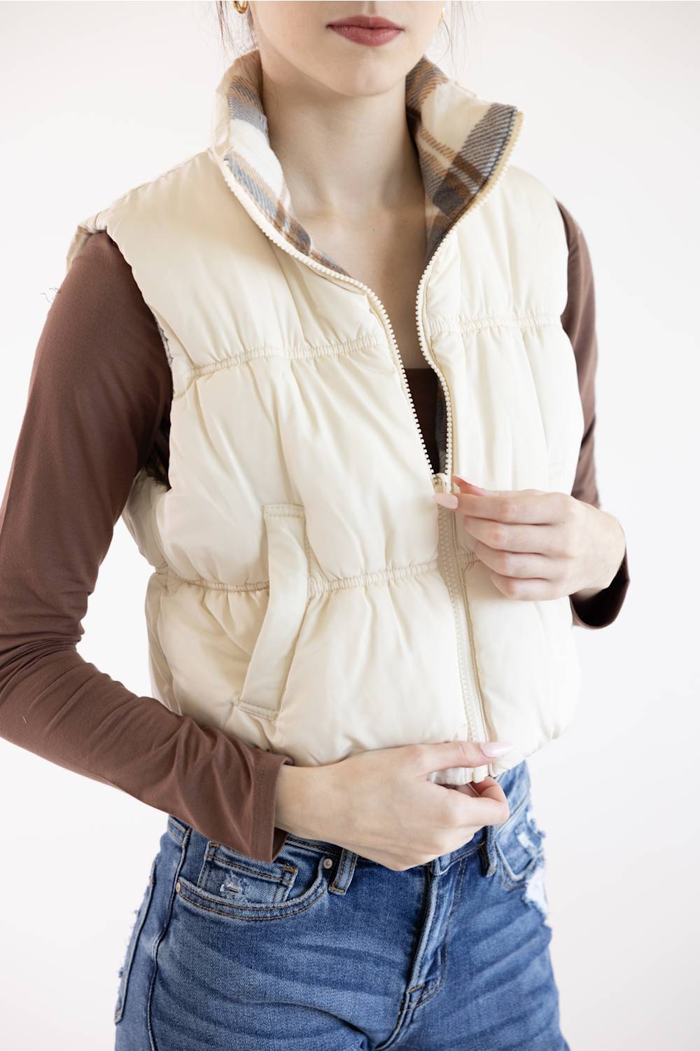 A woman wearing a cream-colored puffer vest over a brown long-sleeve shirt and blue jeans, shown from the chin down against a plain white background.