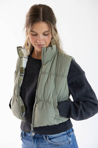 A woman wearing an olive green quilted puffer vest over a dark gray knit sweater and blue jeans, photographed against a clean white studio background.