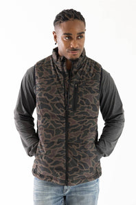 A man wearing a brown and black camouflage patterned zip-up vest over a dark gray long-sleeve shirt and blue jeans, photographed against a plain white background.
