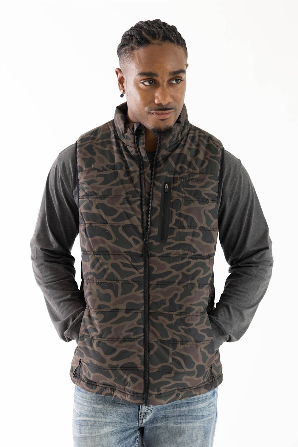 A man wearing a brown and black camouflage patterned zip-up vest over a dark gray long-sleeve shirt and blue jeans, photographed against a plain white background.