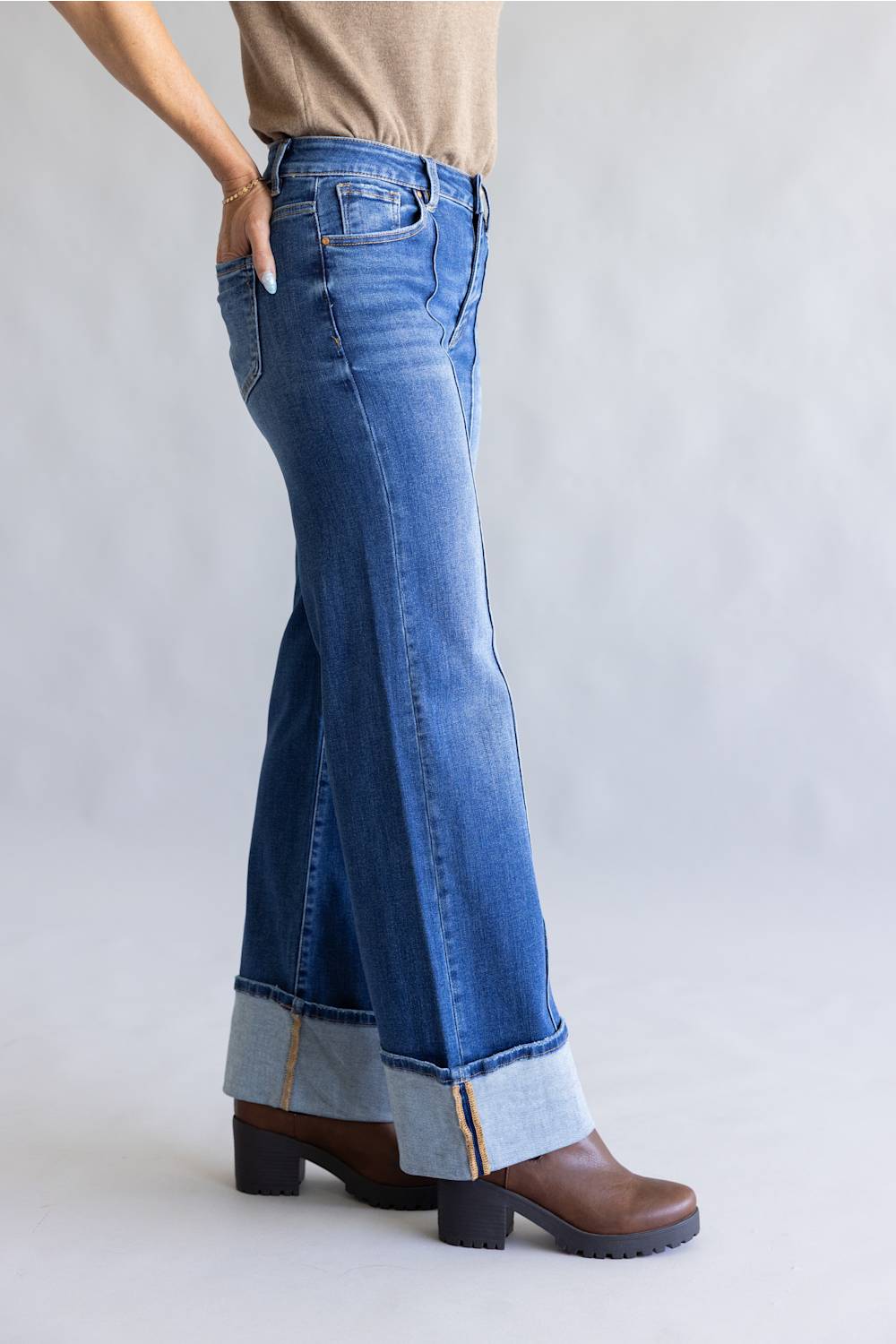 A person wearing medium-wash blue flare jeans with rolled cuffs and brown ankle boots, shown from the waist down against a light gray studio background.