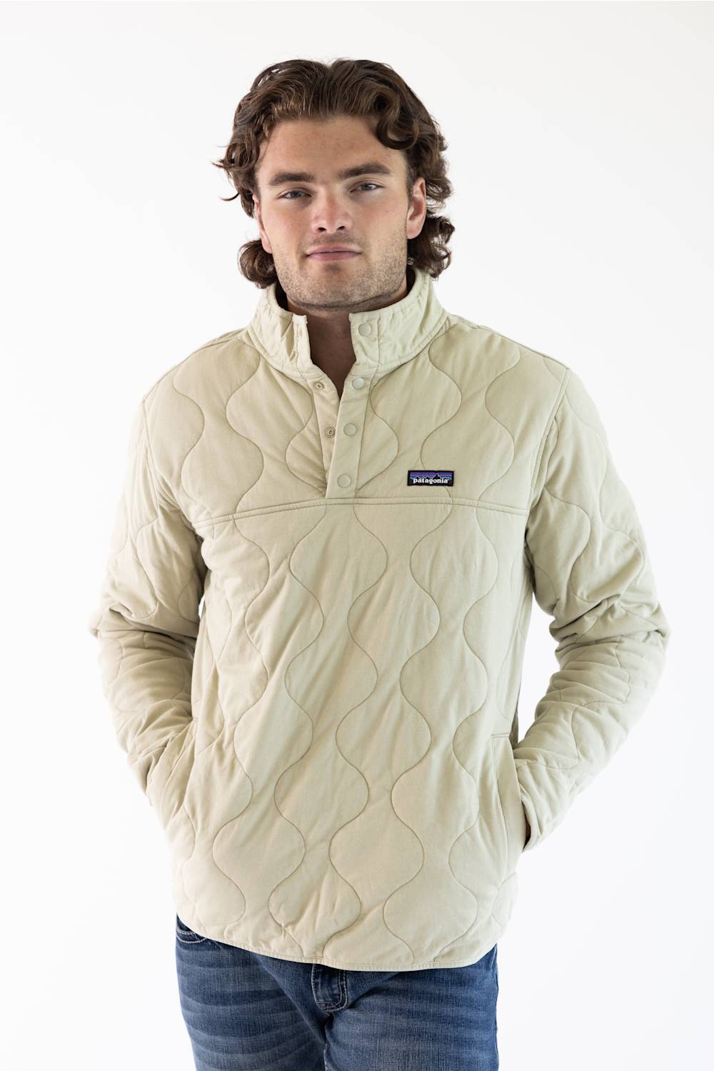 A man wearing an off white Patagonia quilted pullover jacket with a stand collar and zippered chest pocket, paired with blue jeans, against a plain white studio background.
