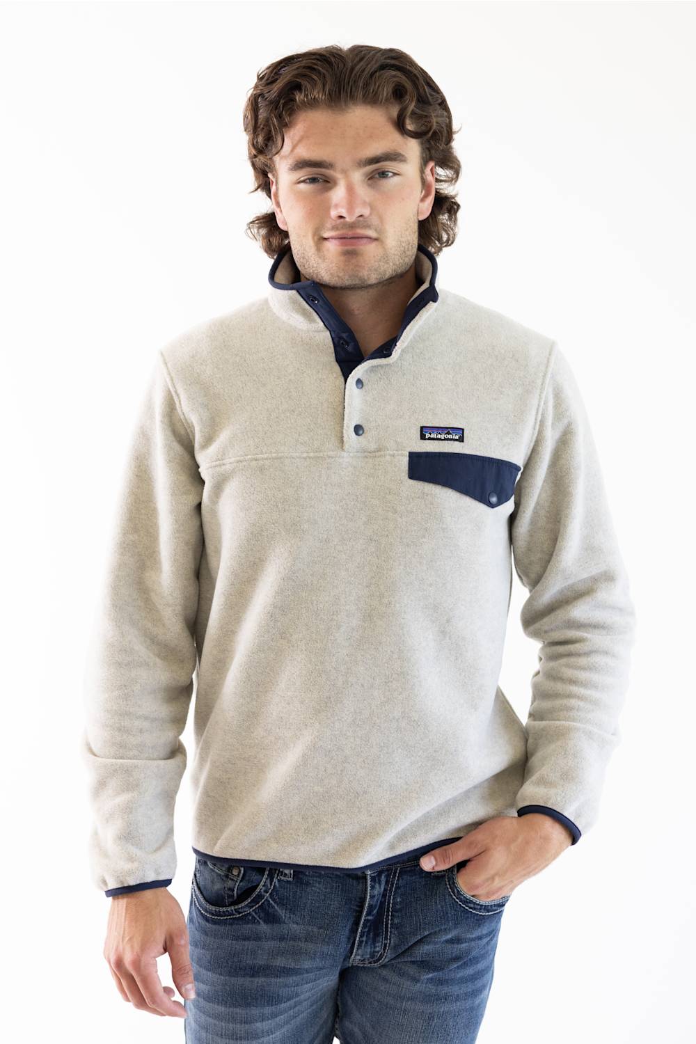 A man wearing a light beige fleece pullover sweater with a navy blue chest pocket and trim, paired with blue jeans, photographed against a plain white studio background.