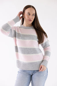 Three-quarter view of a woman wearing the Pink Rose Drop Shoulder Striped Knit Sweater in grey, light pink, and white horizontal stripes with her hand touching her hair, paired with light-wash jeans.