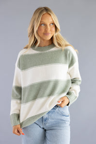 Front three-quarter view of a blonde woman wearing the Pink Rose cream and sage green wide-striped crew neck sweater, styled with light-wash jeans.