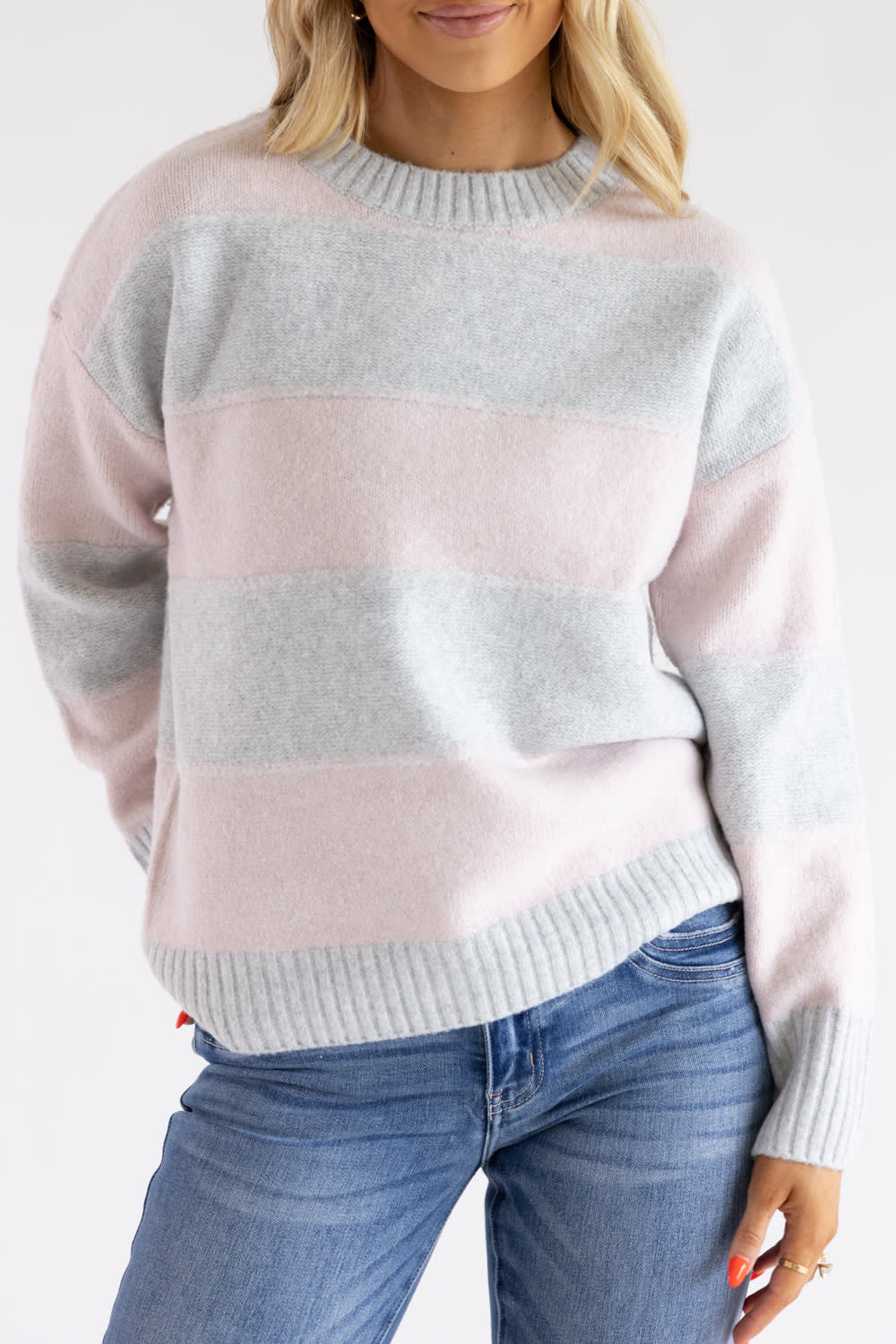Close-up shot of a woman wearing the Pink Rose heather grey and light pink horizontal striped knit crew neck sweater with ribbed collar and hem, styled with blue jeans.