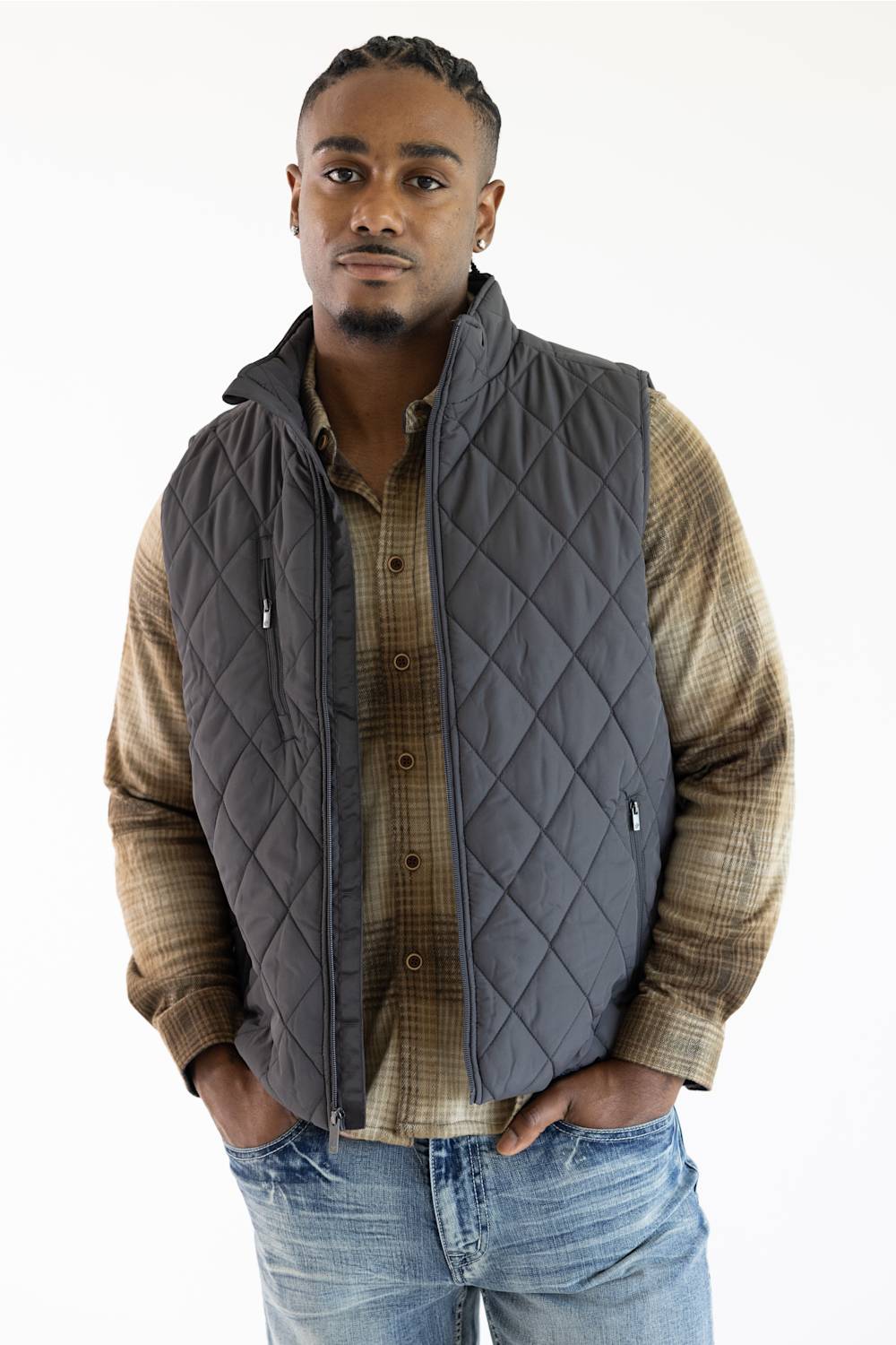 A man wearing a dark gray diamond-quilted zip-front vest over a brown and beige plaid button-up shirt, paired with blue jeans, against a plain white background.