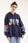 A woman wearing a navy blue hoodie with a colorful patchwork 'JOY' graphic on the chest and matching patchwork details on the sleeves, paired with light blue jeans against a plain white background.