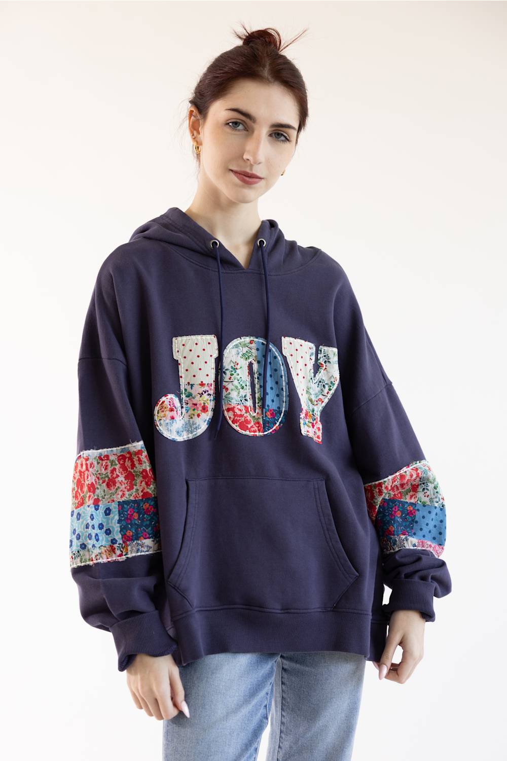 A woman wearing a navy blue hoodie with a colorful patchwork 'JOY' graphic on the chest and matching patchwork details on the sleeves, paired with light blue jeans against a plain white background.