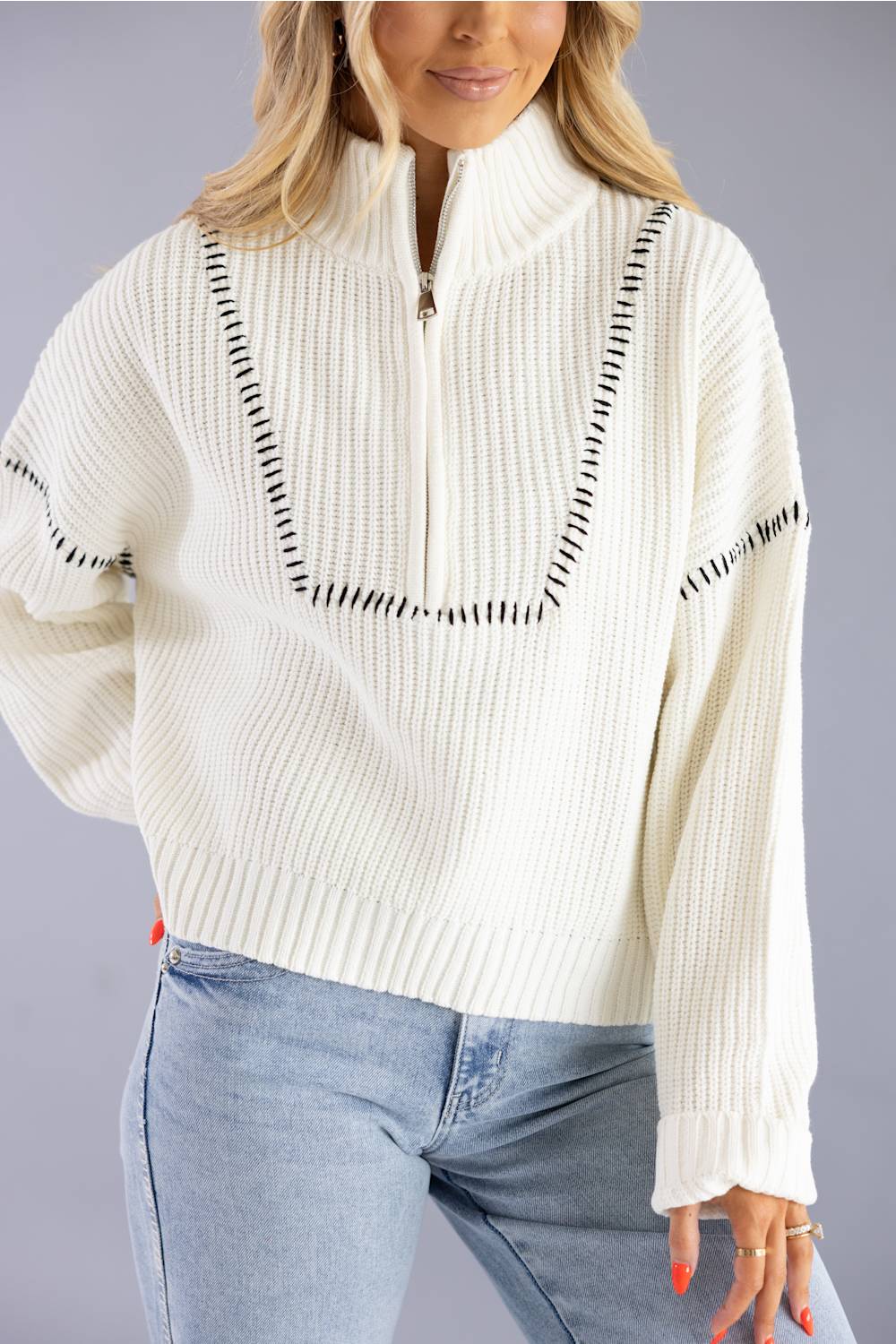A woman wearing a cream ribbed knit half-zip sweater with black dashed line detailing on the chest and sleeves, paired with light blue jeans, against a solid gray studio background.