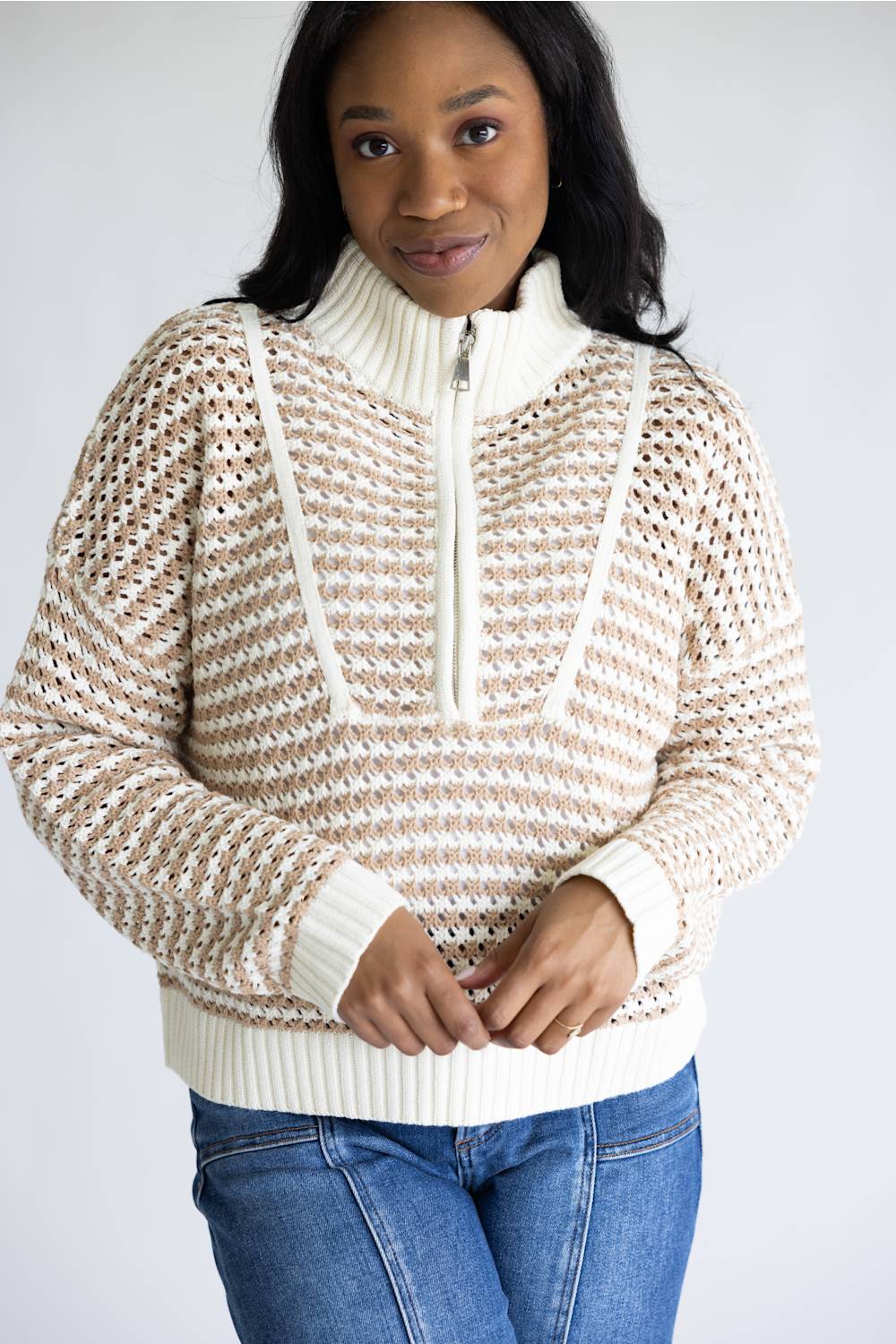 A woman wearing a cream and beige open-knit mock-neck sweater with a half-zip front, paired with blue jeans, against a plain light gray background.