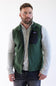 A man wearing a Patagonia green quilted puffer vest over a beige long-sleeve shirt and blue jeans, standing against a plain white background.