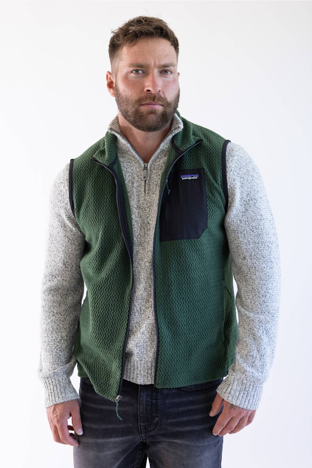 A man wearing a Patagonia green quilted puffer vest over a beige long-sleeve shirt and blue jeans, standing against a plain white background.