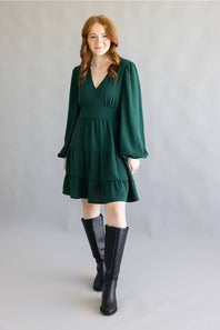 A woman modeling a dark green tiered mini dress with long balloon sleeves and black knee-high boots, standing against a neutral gray studio backdrop.
