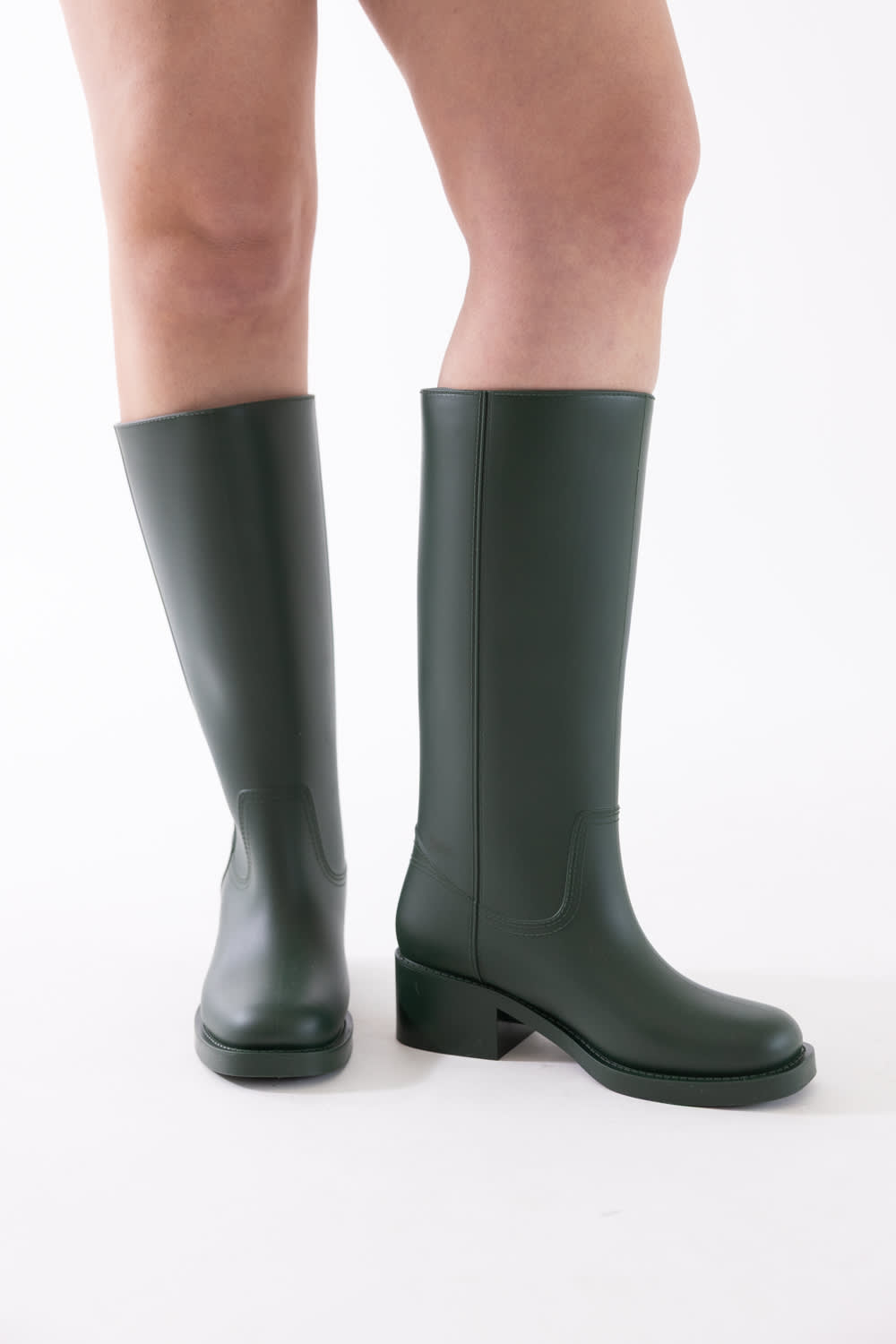 Dark green knee-high rubber rain boots worn on a model's legs, shown from the side against a plain white studio background.
